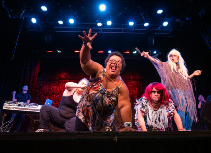 Photo of four performers on stage during the 2019 Disability Drag Show. A woman with Down syndrome is on all fours with her right arm outstretched and grabbing for the audience. Her face is full of joy.