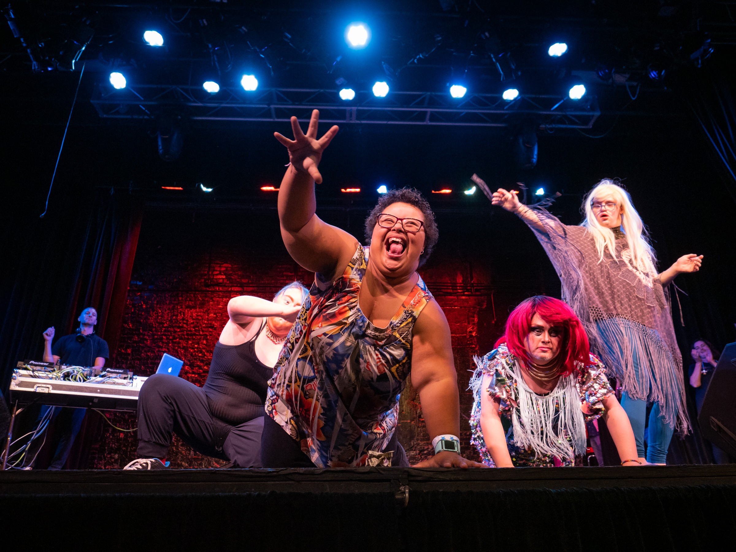 Photo of four performers on stage during the 2019 Disability Drag Show. A woman with Down syndrome is on all fours with her right arm outstretched and grabbing for the audience. Her face is full of joy.