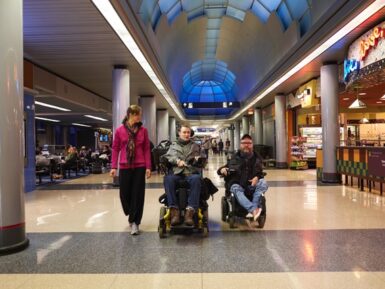 Two men in electronic wheelchairs and a female walking down the corridor of a busy airport.