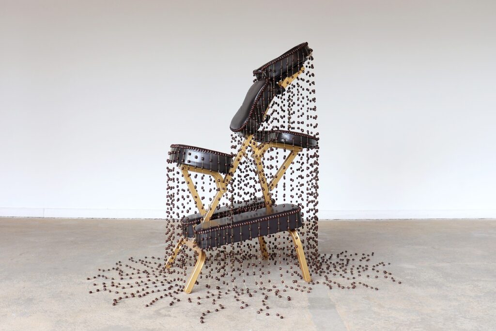 An artistic installation featuring three folding chairs, with parts of each chair appearing to disintegrate into a cascade of small, dark beads that spill onto the gray floor. The surreal and striking composition creates a visual illusion of the chairs unraveling, set against a plain white background in a spacious gallery.
