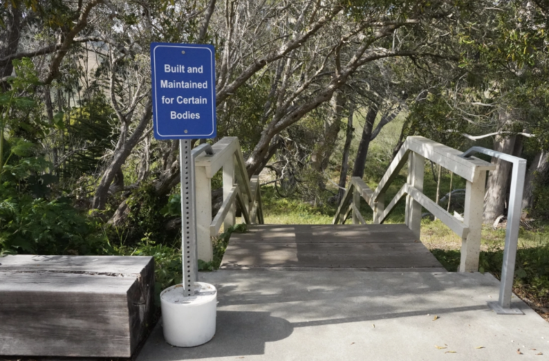 A photograph of a blue industrial sign in front of a concrete staircase leading to a path through a forest. The sign reads “Build and Maintained for Certain Bodies”.