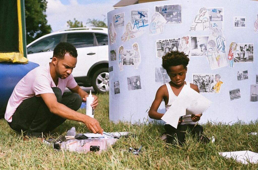 A photograph of two people with dark skin and dark hair sitting in the grass in front of a large poster with printed black and white photos and drawings cut out and glued onto it. The person on the right is young and cutting out a drawing, the other person is holding glue. 