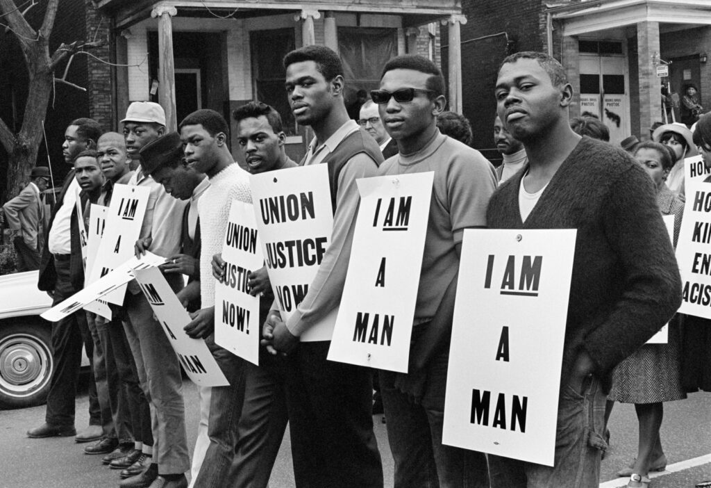 A historical black and white civil rights protest photograph featuring black men with signs that read “I am a man” and “union justice now!”.