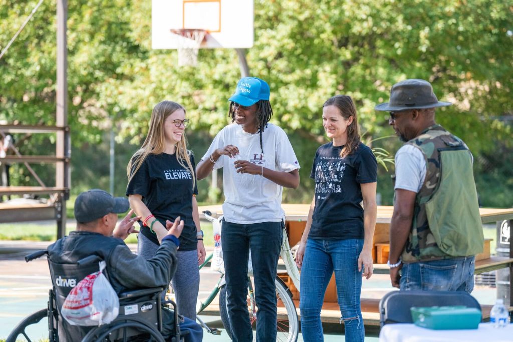 A diverse group of people gathered at an outdoor community event on a sunny day, with a basketball court in the background. The group includes two women in black t-shirts, one wearing a white t-shirt with a blue cap, a man in a camouflage vest and hat, and another man in a wheelchair. They are smiling and engaged in a friendly conversation, creating a warm, inclusive atmosphere.