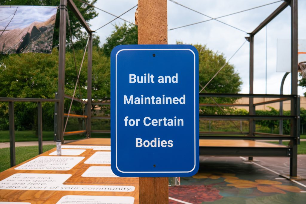 A blue sign mounted on a wooden post in an outdoor setting with the text 'Built and Maintained for Certain Bodies' in white letters. In the background, there is an outdoor display structure with metal frames and informational panels about community, visible under a semi-cloudy sky.