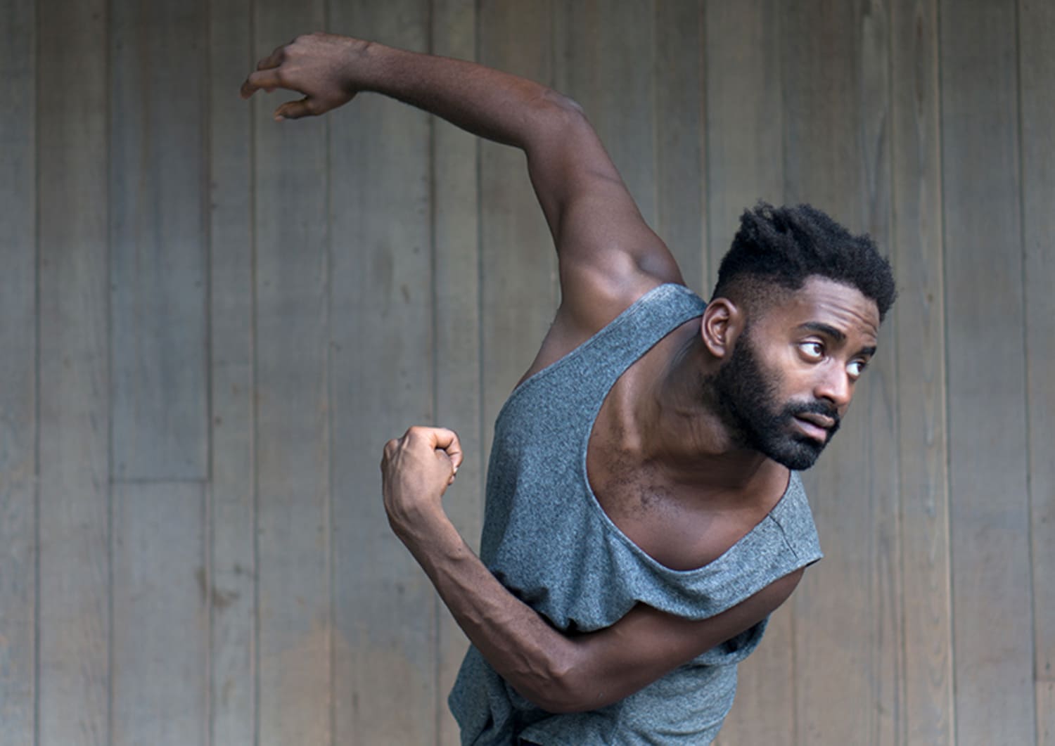 Jerron, a black man with short hair and short facial hair, pictured in motion from the hips up. His left arm is held at a 90 degree angle with his arm close to his body, his fist clenched. His right arm is raised above and behind him, his hand relaxed. He is wearing a graygrey tank top and pictured in front of a wood paneled wall.