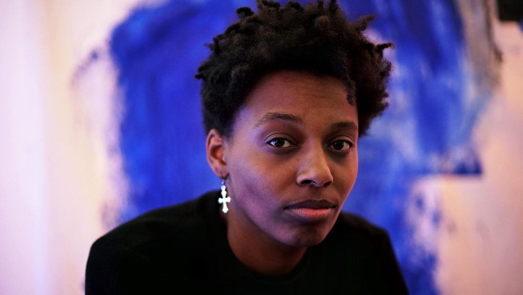 Close-up portrait of a young Black woman with short, natural hair, wearing a black sweater and a silver cross earring. She has a contemplative expression and is looking directly at the camera. The background features an abstract blue painting, adding a vivid contrast to her dark attire and thoughtful demeanor.