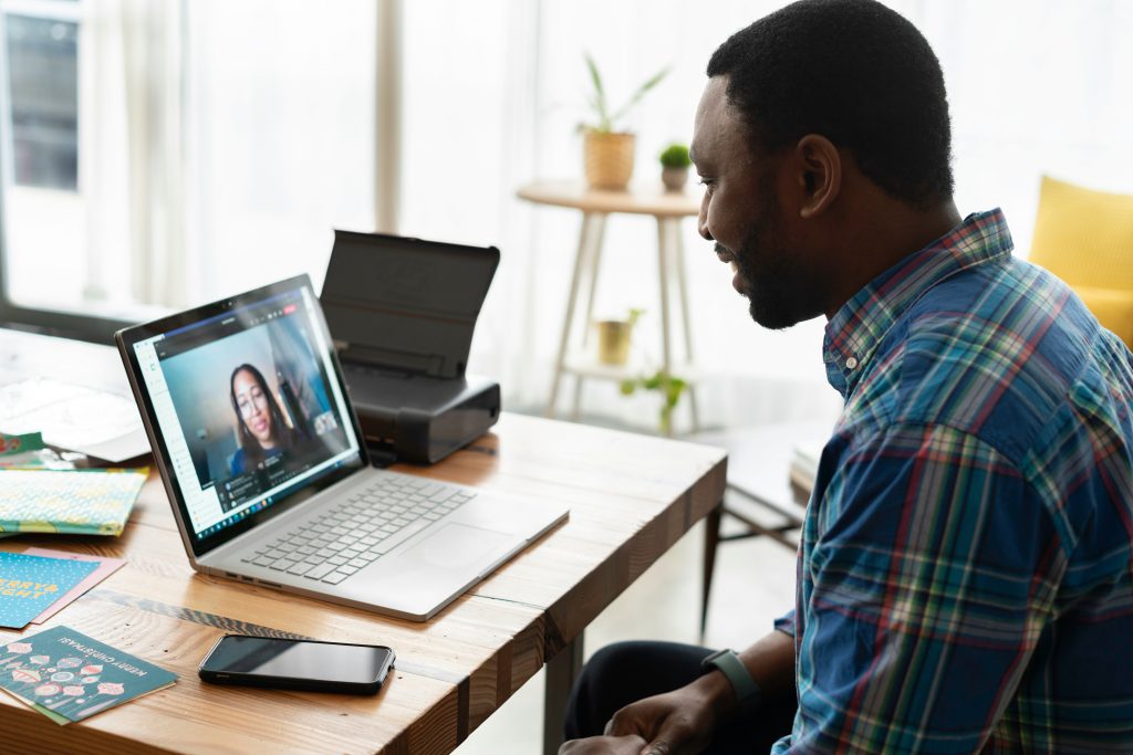 A Black man, seated at a bright and airy home office, is attentively engaged in a video call on his laptop. On the screen, a Black woman appears to be speaking. The desk is neatly organized with another laptop, a printer, some books, and a smartphone. The room is well-lit with natural light streaming in through large windows, and there are potted plants in the background, enhancing the comfortable and productive workspace.