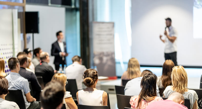 Group of diverse people at a workshop, listening to a speaker who is standing at the front of the room in front of a blank projector screen.